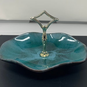 May include: A two-tiered serving dish with a teal and blue glaze. The dish has a wavy rim and a gold-colored handle. The handle is diamond-shaped at the top and connects to the base with a decorative stem. The dish is likely used for serving food or displaying items.
