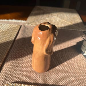 Peut inclure: Un vase en c&eacute;ramique marron en forme de chien avec une finition brillante. Le vase a une ouverture creuse en haut et des d&eacute;tails, dont les oreilles et le museau du chien. L'objet est d&eacute;coratif.