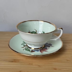 May include: A vintage teacup and saucer set. The cup and saucer are pale green with a gold trim. The cup has a white interior and a handle. The saucer and cup feature a floral design with purple and green accents.