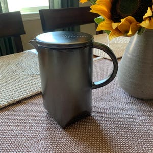 Puede incluir: Una cafetera de prensa francesa gris oscuro con tapa y asa. La cafetera es cilíndrica con base cuadrada. Un jarrón con girasoles está a la derecha.