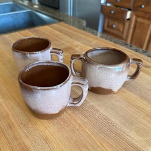 May include: Three ceramic mugs with a brown and cream glaze. The mugs have a rounded shape with a handle. The interior of the mugs is brown, and the exterior has a speckled cream and brown design. The mugs are on a wooden surface.