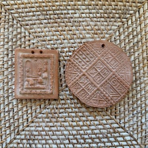 May include: Two terracotta-colored clay ornaments with a matte finish. One is a square plaque featuring a relief of two figures at a table, and the other is a circular disc decorated with a repeating geometric pattern of triangles and lines.