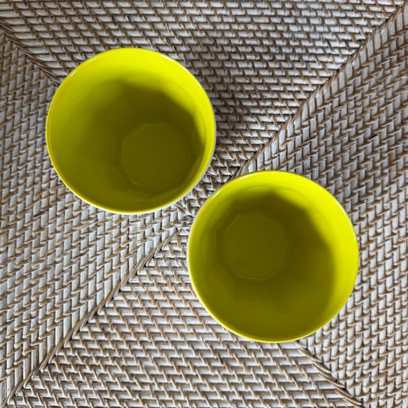 Puede incluir: Dos cuencos de cer&aacute;mica de color amarillo brillante se muestran sobre una superficie tejida de color marr&oacute;n claro. Los cuencos son redondos con un acabado liso y brillante. Los cuencos est&aacute;n vac&iacute;os y listos para ser llenados.