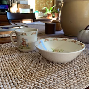 May include: A cream-colored ceramic mug and bowl set. The mug features a handle and a scene of rabbits. The bowl has a textured exterior and a border of running rabbits. Both pieces have a cream-colored background.