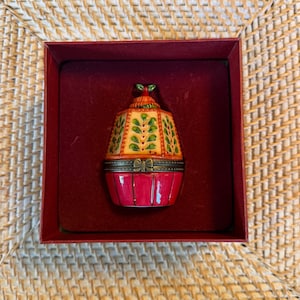 Op de afbeelding: Decoratieve cupcake-vormige sieradendoos. De doos is rood met een gele en groene bovenkant en bevindt zich in een rode doos. De bovenkant heeft een kleine groene strik en een bladerpatroon. De doos is waarschijnlijk gemaakt van porselein of keramiek.