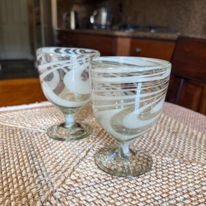 May include: Two clear glass goblets with white and tan swirl designs. The goblets have a short stem and a wide, rounded bowl. They are sitting on a woven placemat. The background includes a kitchen and a wooden chair.