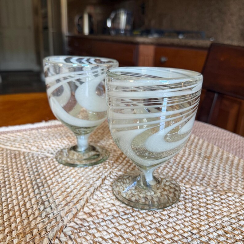 May include: Two clear glass goblets with white and tan swirl designs. The goblets have a short stem and a wide, rounded bowl. They are sitting on a woven placemat. The background includes a kitchen and a wooden chair.