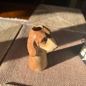 Peut inclure: Un vase en c&eacute;ramique en forme de chien, avec un sch&eacute;ma de couleurs marron et beige. Le vase est con&ccedil;u sous la forme de la t&ecirc;te d'un chien, avec des traits faciaux d&eacute;taill&eacute;s. Le vase est probablement utilis&eacute; pour contenir des fleurs ou comme objet d&eacute;coratif.