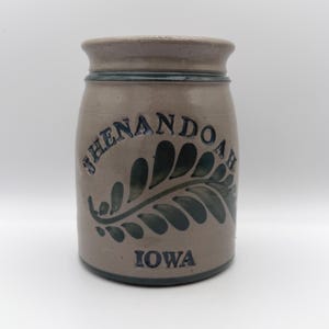 Pode incluir: Um pote de grés cilíndrico com acabamento castanho claro e rebordo azul escuro. A frente apresenta o texto em relevo SHENANDOAH sobre um desenho de folha de feto azul escuro e a palavra IOWA impressa em letras azul escuro na base.