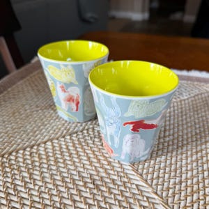 Puede incluir: Dos tazas azul claro con interiores amarillos. Las tazas est&aacute;n decoradas con ilustraciones coloridas de perros. Las tazas est&aacute;n sobre una estera tejida.