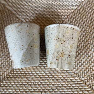 May include: Two handmade ceramic cups with a speckled, textured surface. The cups are off-white with brown and tan speckles. The cups have a slightly tapered shape and a smooth rim. The cups are likely for drinking beverages.