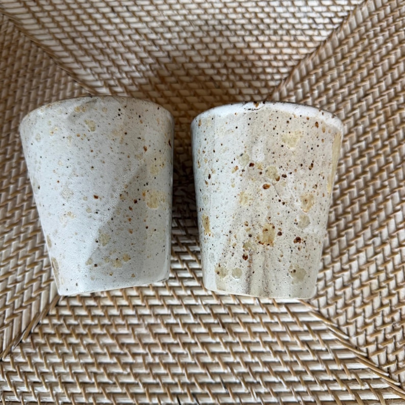 May include: Two handmade ceramic cups with a speckled, textured surface. The cups are off-white with brown and tan speckles. The cups have a slightly tapered shape and a smooth rim. The cups are likely for drinking beverages.