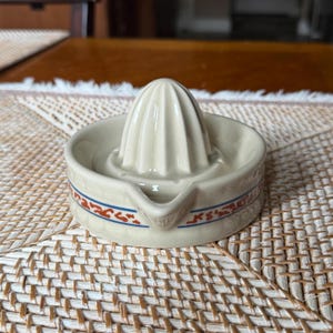 May include: A cream-colored ceramic citrus juicer featuring a central ribbed reamer and a circular basin with a pouring spout. The exterior of the basin is decorated with a horizontal band containing a repeating orange and blue geometric pattern.