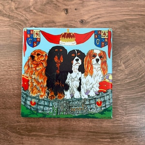 May include: Square ceramic tile featuring four Cavalier King Charles Spaniels. The dogs are depicted in various colors, including black, tan, and white, against a light blue background. A crown and banners with crests are above the dogs.