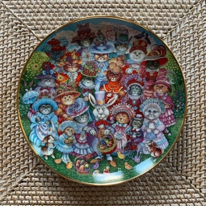 May include: A decorative plate featuring a whimsical illustration of cats dressed in elaborate hats and clothing, gathered in a garden setting. The plate has a gold rim and a colorful design, with a rabbit in the center. The theme is Easter.