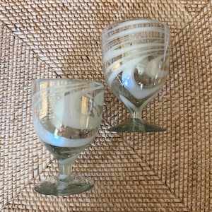 May include: Two clear glass goblets with white swirling designs. The goblets have a stem and a wide base. The glass has a slight green tint. The goblets are on a woven, tan surface.