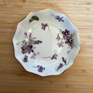 May include: A small, white, scalloped-edge porcelain dish featuring a delicate hand-painted pattern of purple violet flowers and green leaves scattered across the interior surface, finished with a thin, reflective gold trim along the rim.