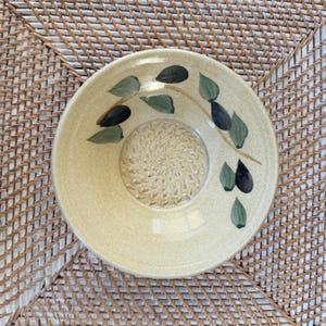 May include: A ceramic garlic grater plate with a light yellow base. The plate features a central textured grating surface and is decorated with a painted branch design with green and black leaves. The plate is round and has a slightly raised edge.