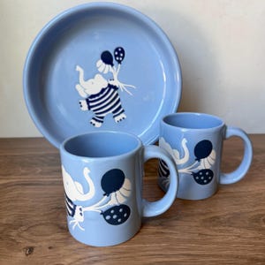 May include: A light blue ceramic dinnerware set consisting of one round plate and two mugs. Each piece features a white elephant graphic wearing a dark blue striped shirt and holding a cluster of dark blue and white balloons with polka dots.