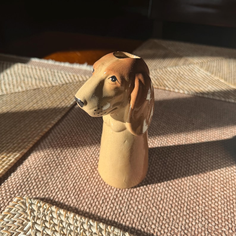 Peut inclure: Figurine de chien teckel en c&eacute;ramique, avec une t&ecirc;te et des oreilles marron et un corps beige. Le chien a un nez noir et regarde vers la gauche. L'objet est d&eacute;coratif, peut-&ecirc;tre un vase ou un porte-crayons.