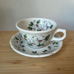 Puede incluir: Un juego de taza de té y plato a juego de cerámica blanca con un patrón pintado de flores de cornejo blancas con hojas verdes y centros de color marrón rojizo. La taza tiene un borde festoneado y un asa curva, al igual que el plato.
