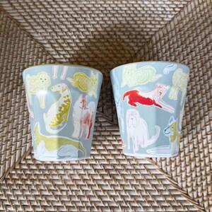 Puede incluir: Dos vasos de cer&aacute;mica azul claro con un patr&oacute;n repetido de varias razas de perros en amarillo, rojo y blanco. Los vasos son cil&iacute;ndricos y est&aacute;n sobre una superficie tejida.