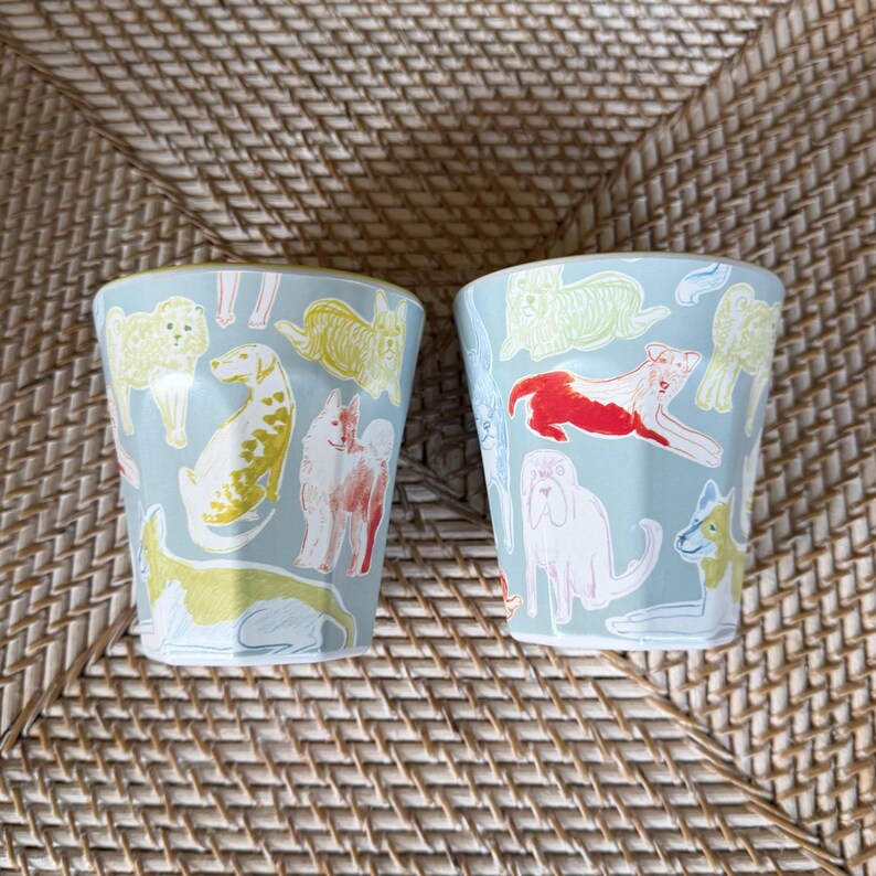 Puede incluir: Dos vasos de cer&aacute;mica azul claro con un patr&oacute;n repetido de varias razas de perros en amarillo, rojo y blanco. Los vasos son cil&iacute;ndricos y est&aacute;n sobre una superficie tejida.
