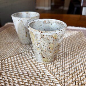 May include: Two handmade ceramic cups with a speckled, off-white glaze and tan accents. The cups have a tapered shape and are set on a woven mat. The cups are likely for drinking beverages.