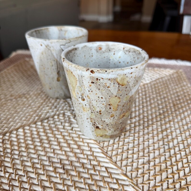 May include: Two handmade ceramic cups with a speckled, off-white glaze and tan accents. The cups have a tapered shape and are set on a woven mat. The cups are likely for drinking beverages.