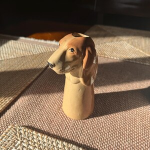 Peut inclure: Figurine de chien teckel en c&eacute;ramique, avec une t&ecirc;te et des oreilles marron et un corps beige. Le chien a un nez noir et regarde vers la gauche. L'objet est d&eacute;coratif, peut-&ecirc;tre un vase ou un porte-crayons.