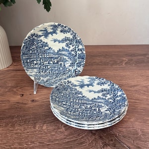 Può includere: Un set di piatti rotondi in ceramica con un motivo decorativo blu e bianco. Il design raffigura una dettagliata scena pastorale con una carrozza trainata da cavalli, edifici a graticcio, alberi e un cielo nuvoloso. I piatti hanno bordi smerlati.