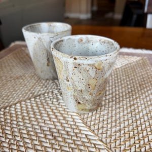 May include: Two handmade ceramic cups with a speckled, textured surface. The cups have a light beige base color with brown and yellow accents. The cups are placed on a woven mat, suggesting a rustic or artisanal theme.