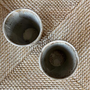 May include: Two speckled ceramic cups with a gray and brown color scheme. The cups are cylindrical with a slightly flared rim. The interior of the cups is a lighter shade of gray with brown speckles. The cups are sitting on a woven wicker surface.