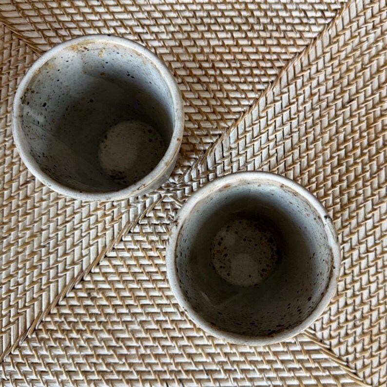 May include: Two speckled ceramic cups with a gray and brown color scheme. The cups are cylindrical with a slightly flared rim. The interior of the cups is a lighter shade of gray with brown speckles. The cups are sitting on a woven wicker surface.