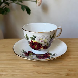 May include: A white porcelain teacup and saucer set featuring a floral pattern with deep red, white, and green petals and leaves. The rim of both the cup and the saucer is finished with a thin gold trim, and the cup has a curved handle.
