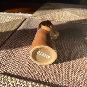 Peut inclure: Figurine en c&eacute;ramique marron avec une &eacute;tiquette sur le dessous indiquant "Made in China / Fabriqu&eacute; en Chine". La figurine est pos&eacute;e sur une surface textur&eacute;e brun-rouge.