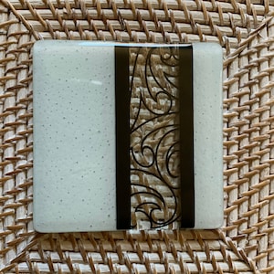 May include: Square, light-colored glass coaster with a dark, vertical, decorative design. The design features a black, ornate, swirling pattern. The coaster has a textured surface and is sitting on a woven, brown surface.