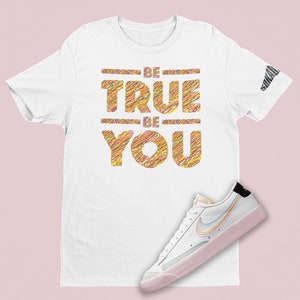 Puede incluir: Camiseta blanca con el texto "BE TRUE BE YOU" en letras coloridas y texturizadas. Una zapatilla blanca y rosa con un swoosh arcoíris se encuentra en la parte inferior derecha de la imagen.