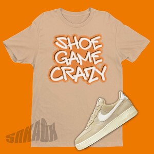 May include: A tan t-shirt with the text "Shoe Game Crazy" in white and orange lettering. A pair of tan suede trainers with a white swoosh logo are shown next to the t-shirt.