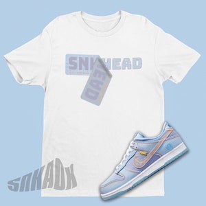 May include: White t-shirt with the text "SNEAKHEAD" in a blue and orange grid pattern. A blue and white Nike Dunk Low sneaker with a light blue swoosh and a yellow tag.