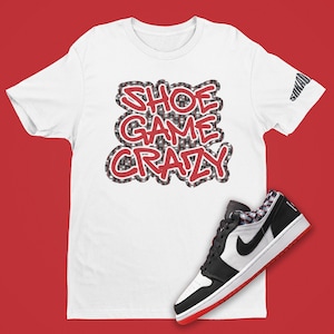 May include: White t-shirt with the text "Shoe Game Crazy" in red and black letters with a checkered pattern. A black and white sneaker with red accents is shown below the shirt.
