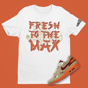 May include: White t-shirt with orange text that says "FRESH TO THE MAX" with a graphic of a dragon. A pair of tan, orange, and red Nike Air Max 1 sneakers are shown below the t-shirt.