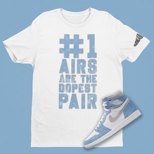 May include: White t-shirt with the text "#1 AIRS ARE THE DOPEST PAIR" in light blue. A light blue and white high-top sneaker is next to the shirt. The sleeve of the shirt has the word "SAKAD" printed on it.