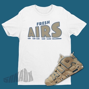 May include: A white t-shirt with the text "FRESH AIRS" in brown and blue lettering. A pair of tan and blue Nike Air More Uptempo sneakers are shown below the shirt.