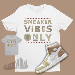 May include: White t-shirt with the text "SNEAKER VIBES ONLY" in brown and beige letters. A pair of white, beige, and brown trainers are on the bottom right of the shirt.