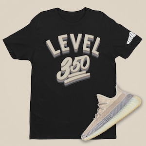 Puede incluir: Camiseta negra con el texto "LEVEL 350" en estilo 3D. Un par de zapatillas beige y blancas se encuentran en la parte inferior derecha de la imagen.