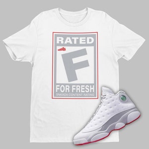 May include: White t-shirt with a graphic of a red and white "Rated F for Fresh" logo. A pair of white and grey sneakers with red accents are shown below the t-shirt.