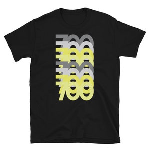 Op de afbeelding: Zwart T-shirt met een grafisch ontwerp van het getal 700 in geel en grijs. Het getal is gelaagd en gestapeld op zichzelf.