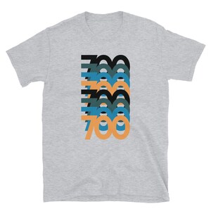Peut inclure: Un t-shirt gris chin&eacute; avec un motif graphique repr&eacute;sentant le nombre 700 dans un design superpos&eacute;, en couches et color&eacute;. Les chiffres sont dans des tons de noir, bleu sarcelle, bleu et orange.