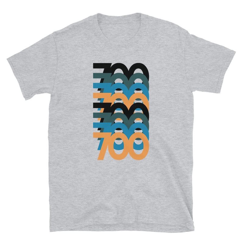 Peut inclure: Un t-shirt gris chin&eacute; avec un motif graphique repr&eacute;sentant le nombre 700 dans un design superpos&eacute;, en couches et color&eacute;. Les chiffres sont dans des tons de noir, bleu sarcelle, bleu et orange.